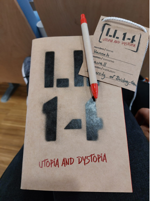 Brown paper notebook, paper lanyard and pen with a red tip. The notebook and the lanyard have a graffiti-style logo that reads 'LL 14' in black and 'Utopia and Dystopia' in red letters. The lanyard also has my name (Hannah Sawall) and my affiliation 'University of Duisburg-Essen'. You can see the back of seats in a lecture hall and the floor in the background.